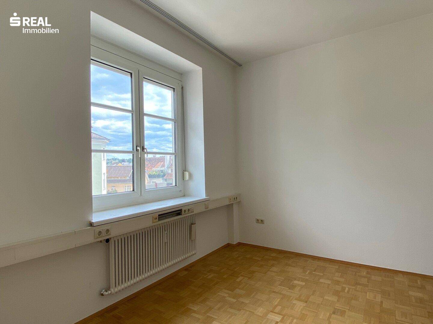 Einzelner Büroraum mit großem Fenster, Heizkörper und Holzparkettboden, gut gepflegt.