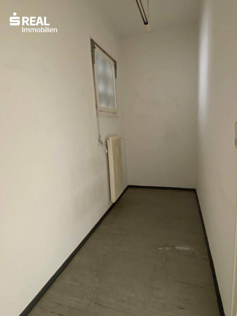 Schmaler Abstellraum mit Betonboden und kleinem Fenster, geeignet für Lagerzwecke.