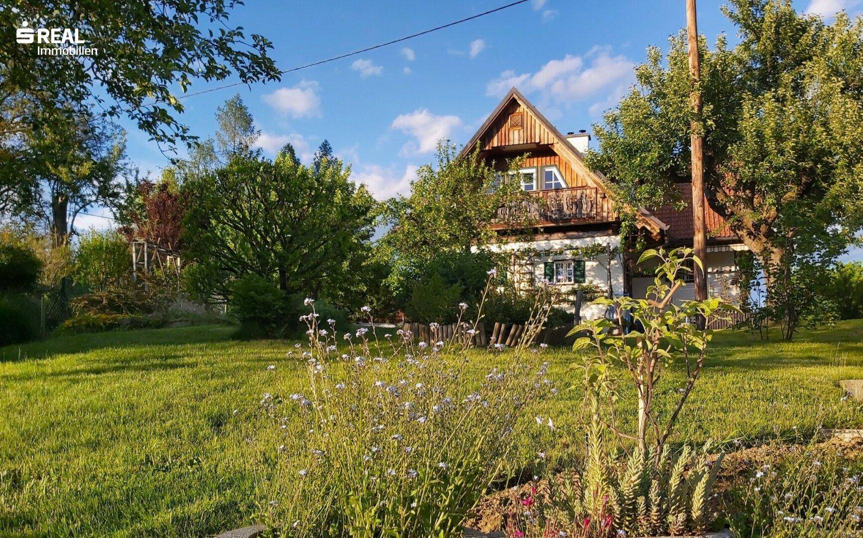 Idyllisches Landhaus umgeben von üppigem Grün und blühenden Pflanzen unter blauem Himmel.