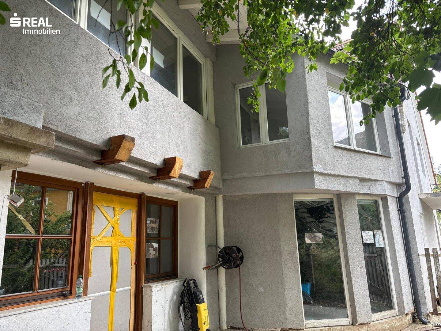 Detailansicht der Hausfassade mit gelber Tür und großen Fenstern, umgeben von Bäumen.
