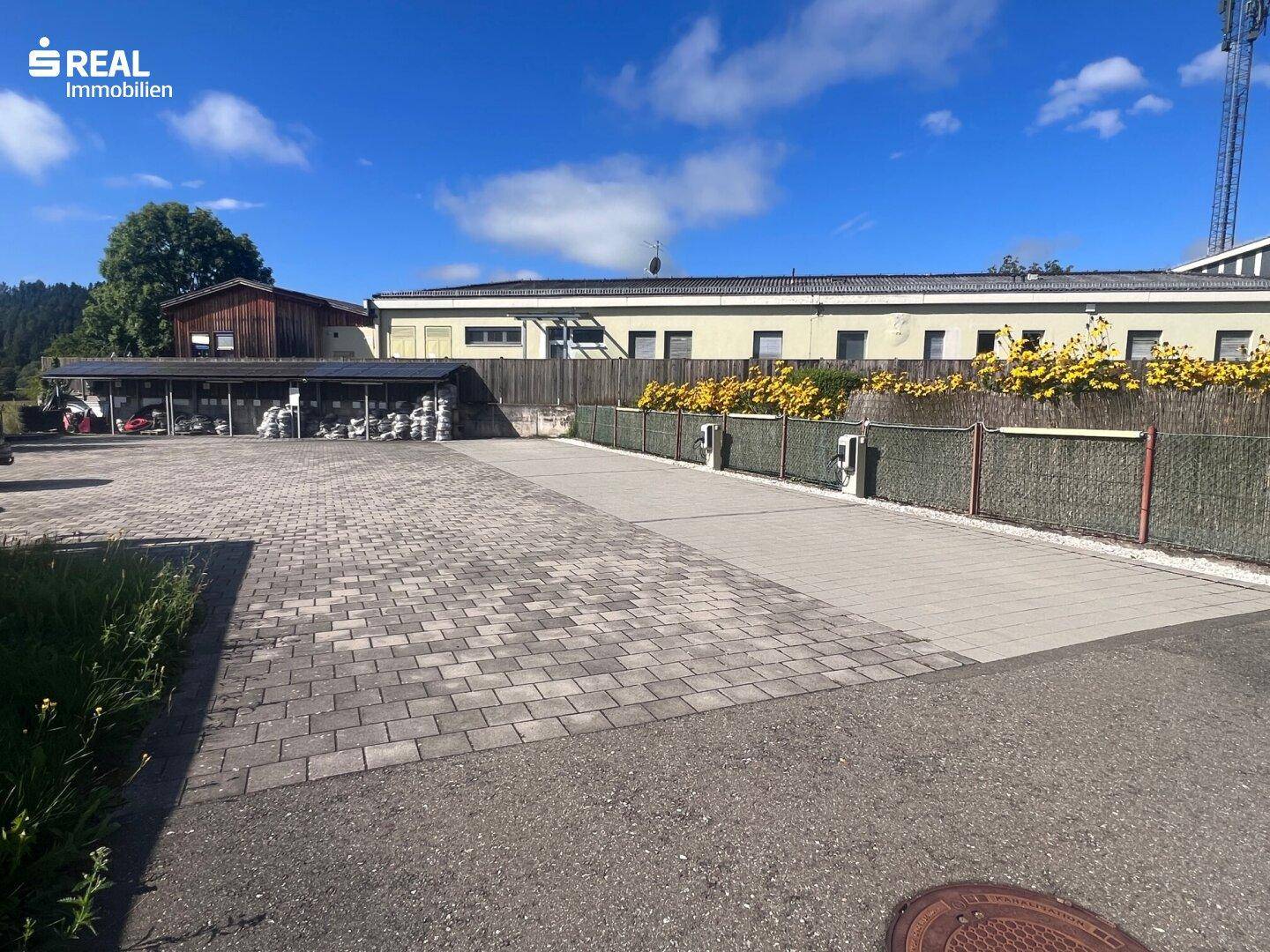 Großer gepflasterter Parkplatz mit Ladestationen für Elektrofahrzeuge und grüner Hecke.