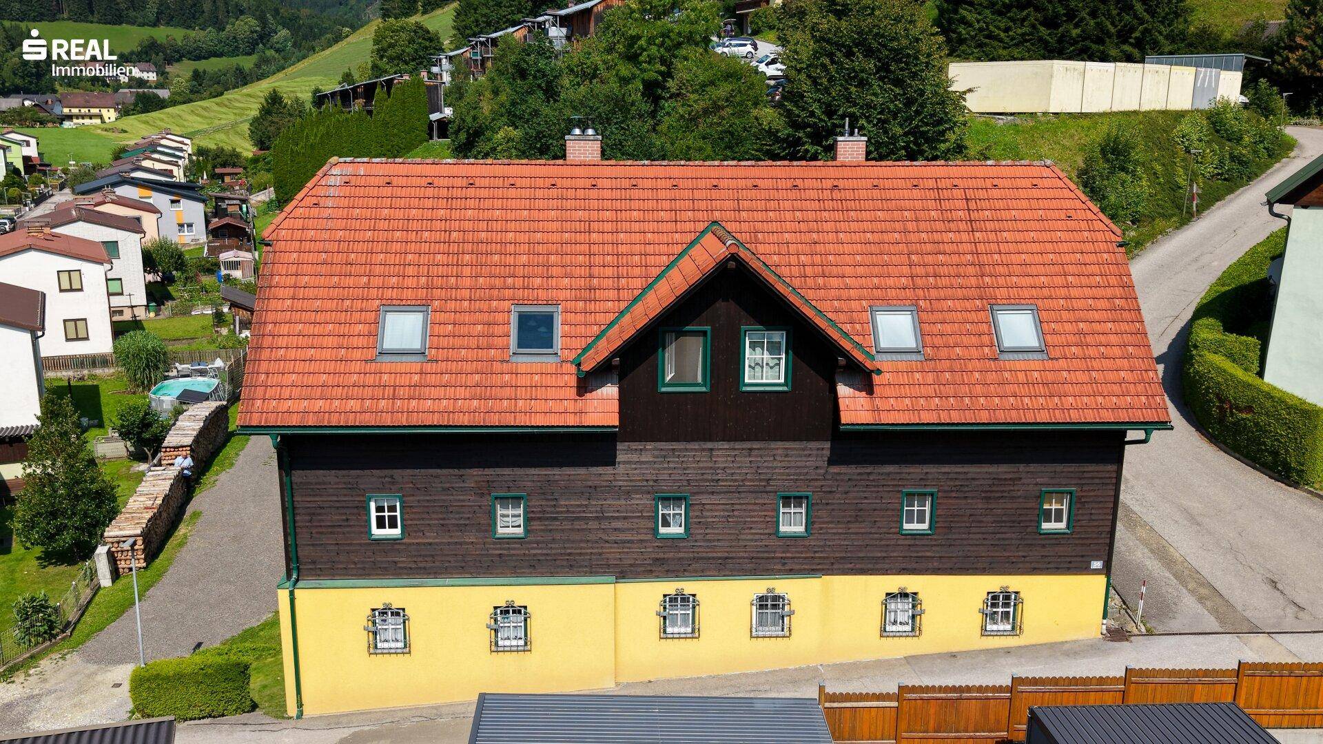 Draufsicht auf das Haus mit markantem roten Ziegeldach, Gauben und Dachfenstern in ländlicher Umgebung.