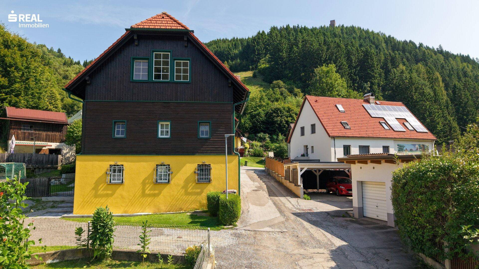 Vorderansicht des Hauses mit gelbem Sockel und dunkler Holzfassade, umgeben von ländlicher Bebauung und Natur.