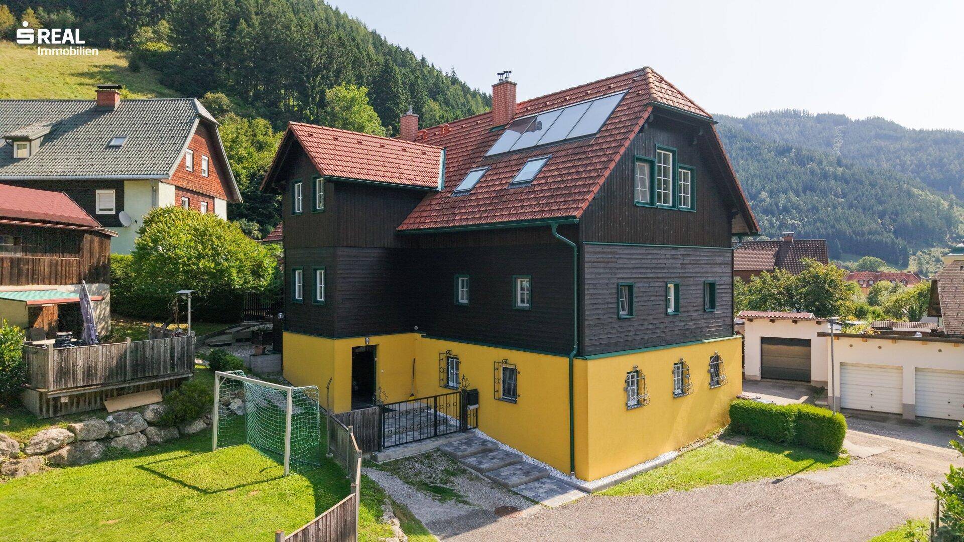 Ansicht des Hauses mit gelbem Sockel, dunkler Holzfassade und rotem Ziegeldach, umgeben von grüner Landschaft.