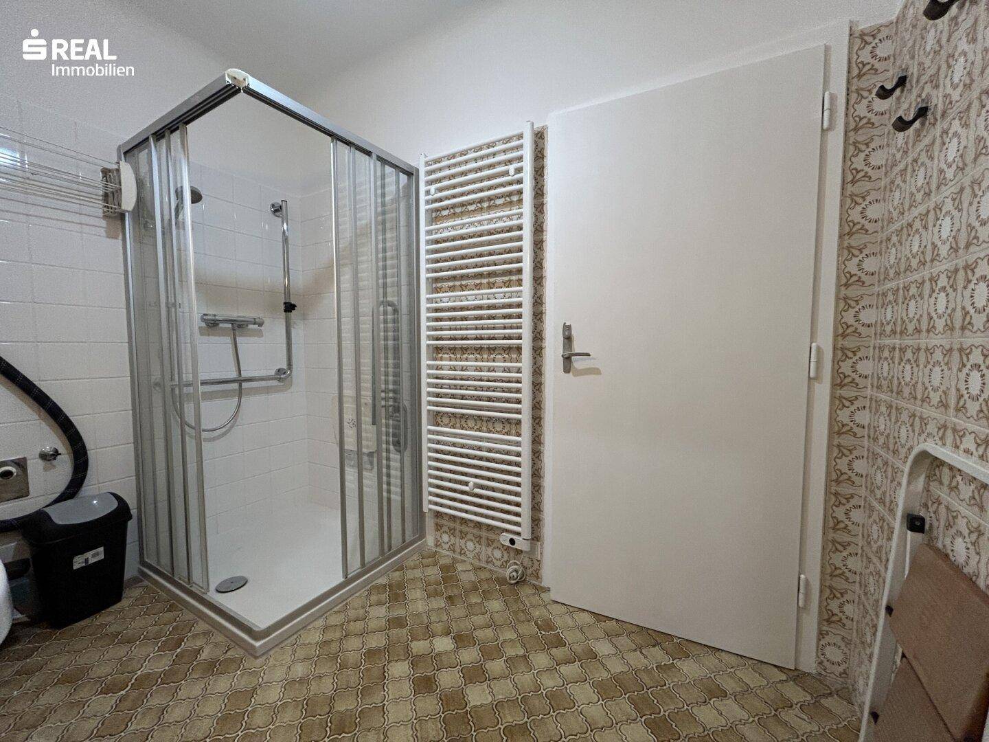 Badezimmer mit Duschkabine, Handtuchheizkörper und gemusterten Fliesen im 80er-Jahre-Stil.