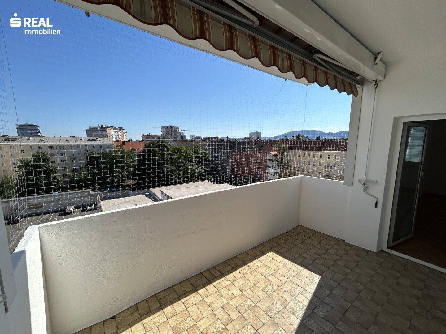 Heller Balkon mit Stadtblick, Fliesenboden und Schutznetz unter einer Markise.