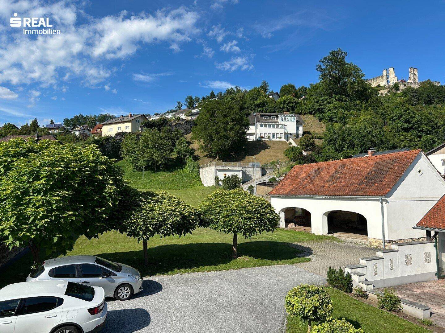 Außenbereich mit Parkplätzen und einem Nebengebäude, umgeben von grünen Hügeln und Ruinen.