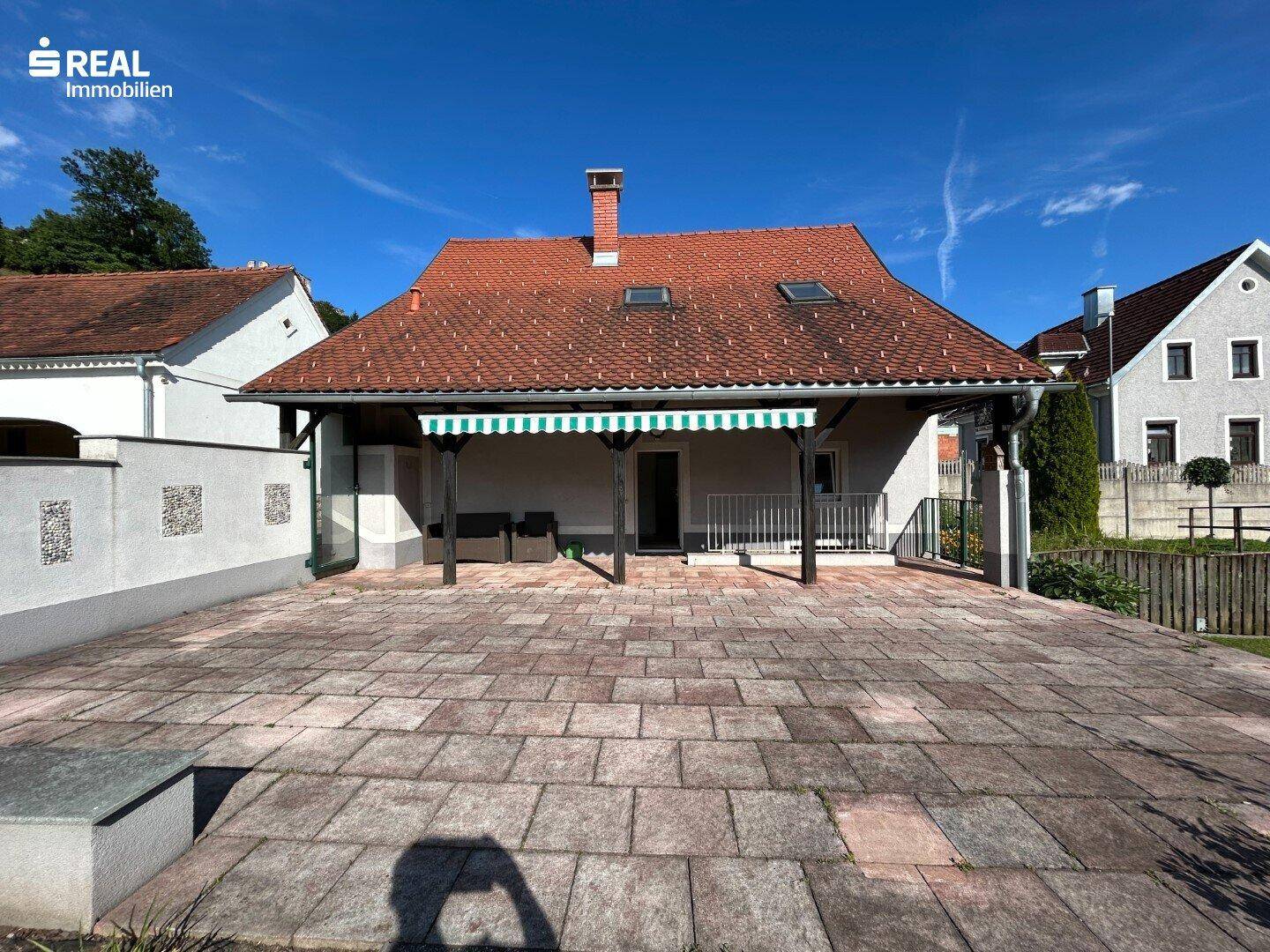 Weitläufige gepflasterte Terrasse mit Markise und Blick auf die grüne Umgebung.