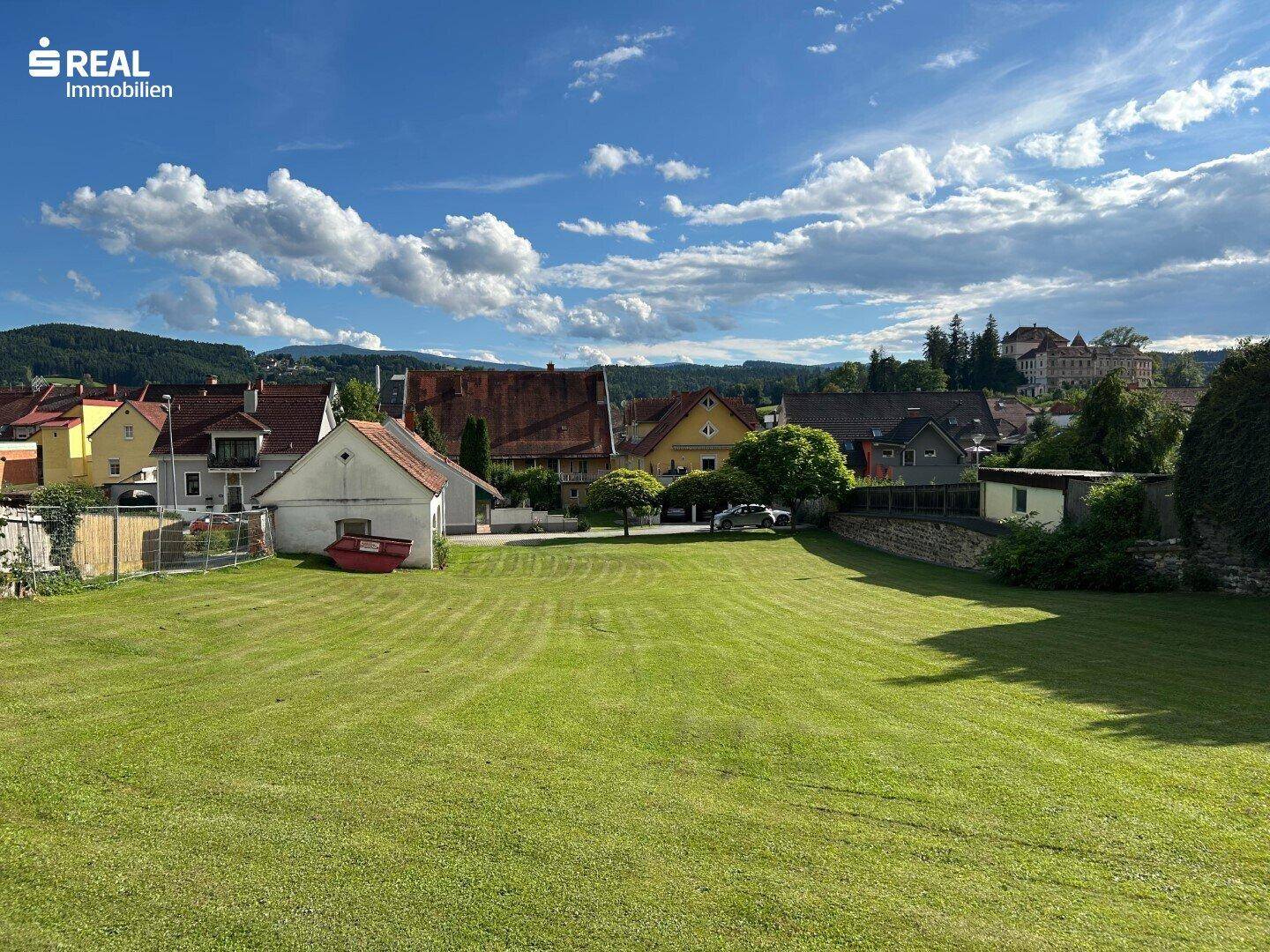 Weitläufige Grünfläche mit Blick auf die umliegende Landschaft und entfernte Häuser unter blauem Himmel.