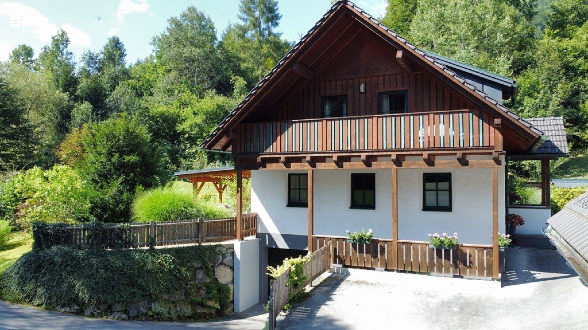 Idyllisches Landhaus mit traditionellem Holzbalkon und Zufahrt zur Garage, eingebettet in grüne Landschaft.