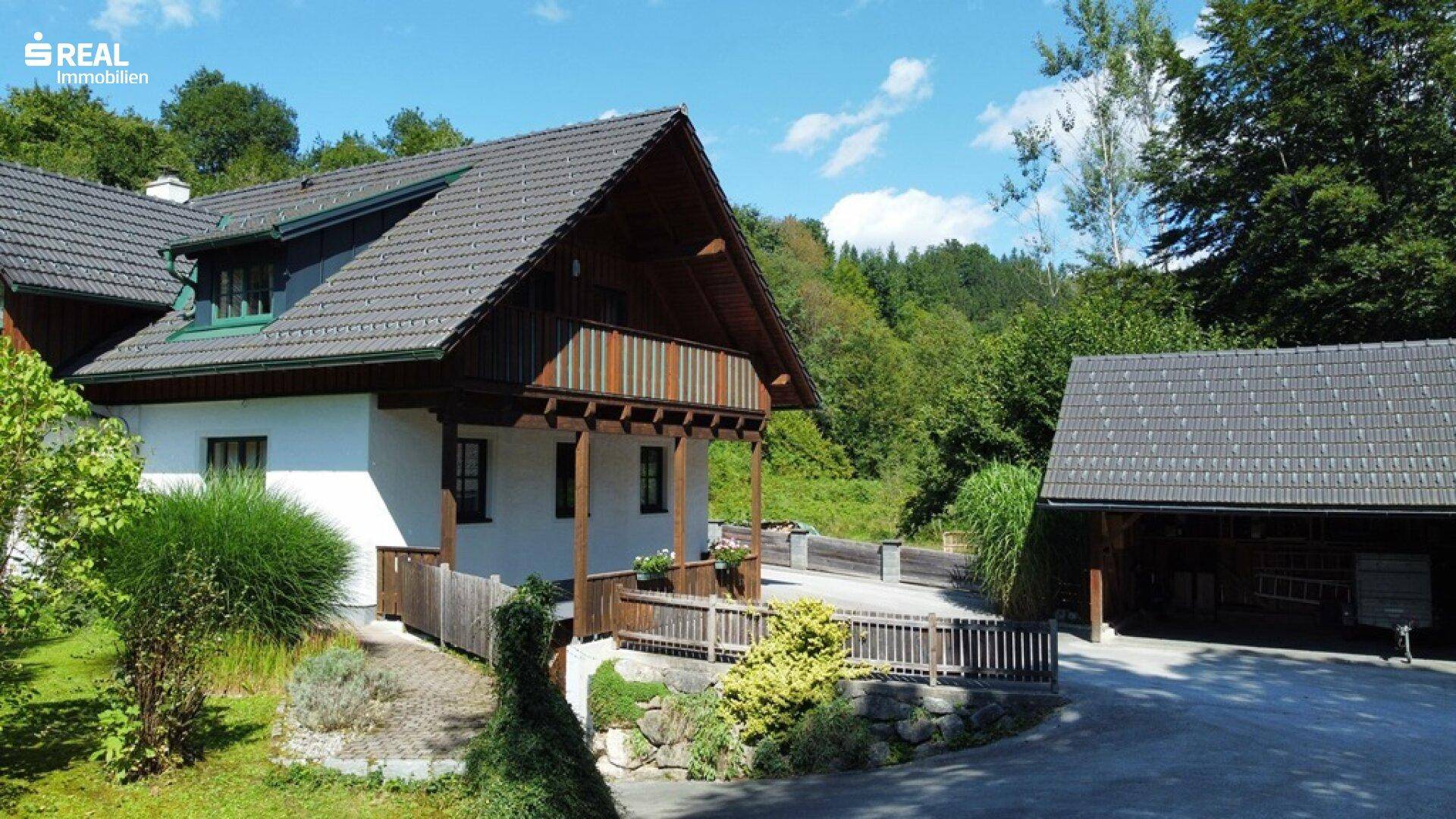 Seitliche Ansicht des Hauses mit Holzbalkon und einem überdachten Carport, umgeben von viel Grün.