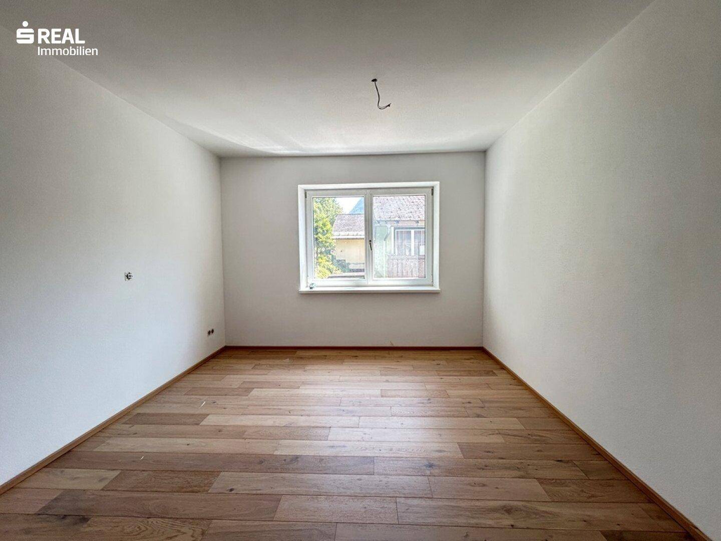 Leeres Zimmer mit hellen Wänden, Holzboden und einem Fenster, das Tageslicht hereinlässt.
