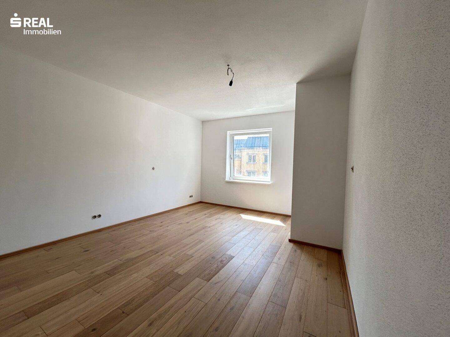Helles, unmöbliertes Zimmer mit Holzboden und einem Fenster, das eine gute Belichtung ermöglicht.