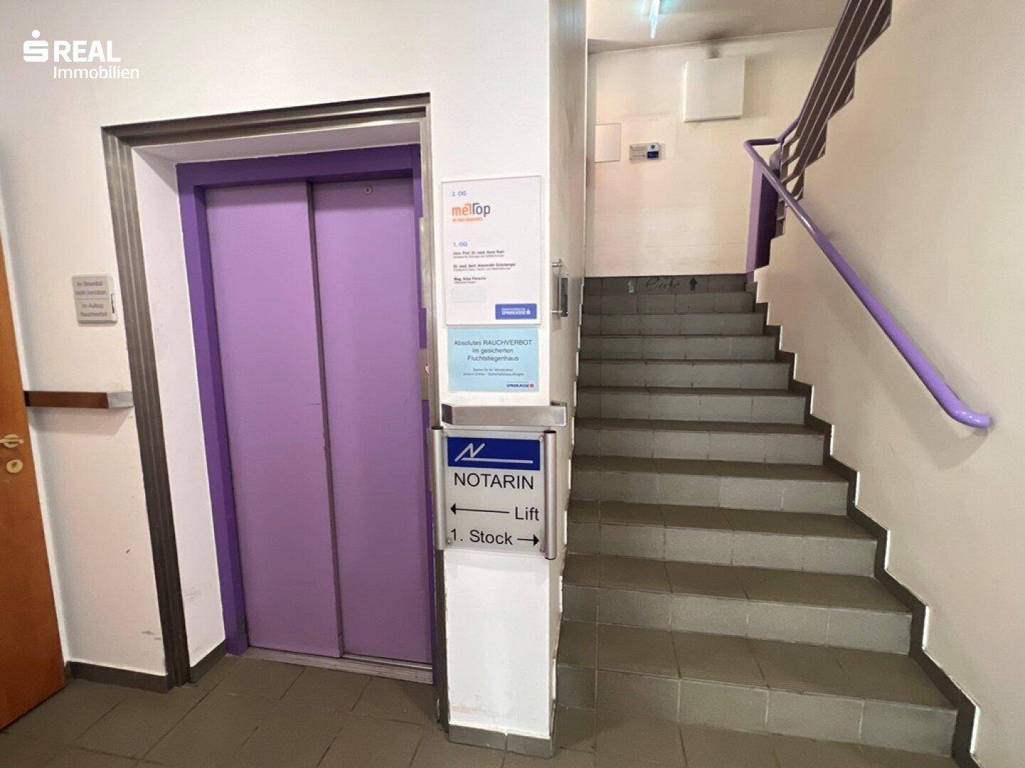 Treppenhaus und Aufzug in lila Farbe, mit Hinweisschildern zum ersten Stock.