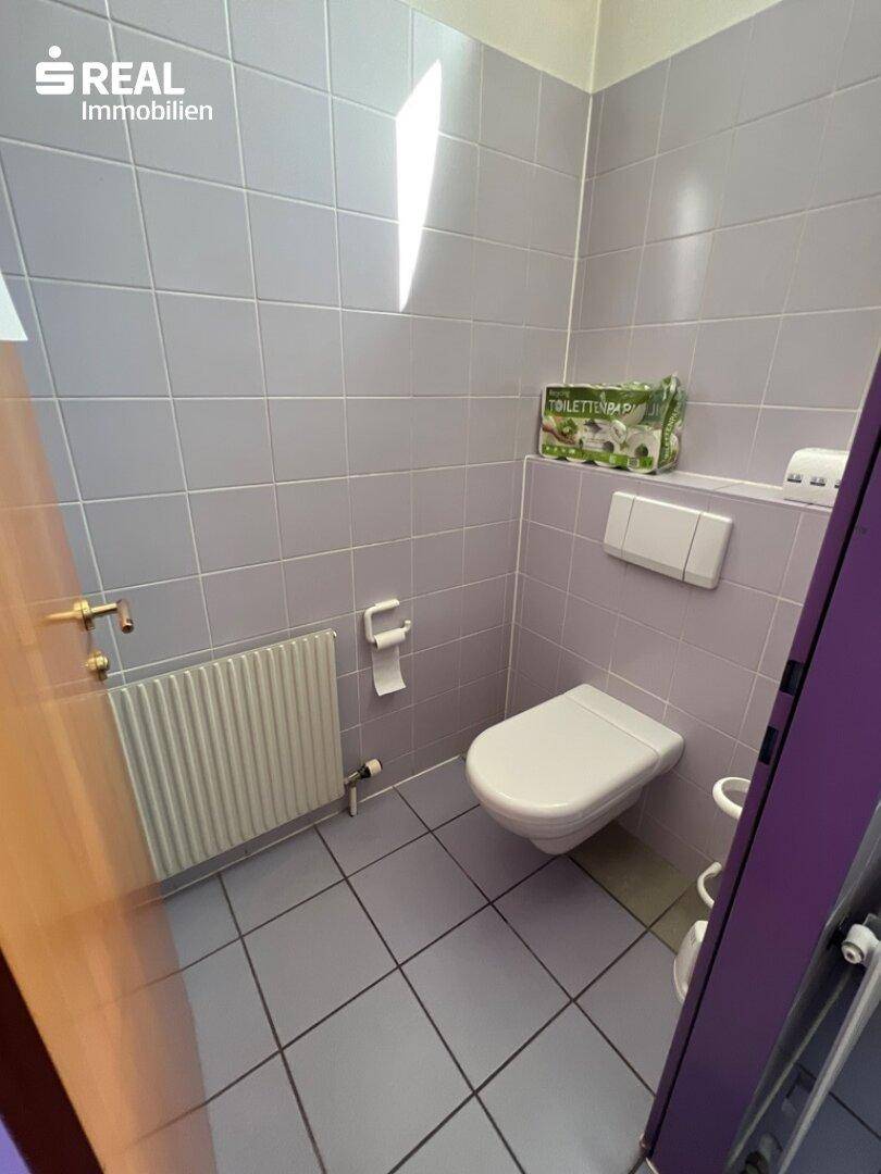 Einfacher Toilettenraum mit wandhängendem WC, Heizkörper und lila Wandfliesen.