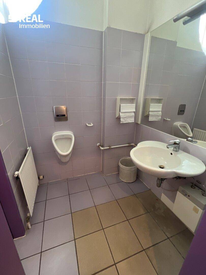 Funktionaler Sanitärbereich mit Urinal, Waschbecken und lila Fliesen, ausgestattet mit einem Heizkörper.