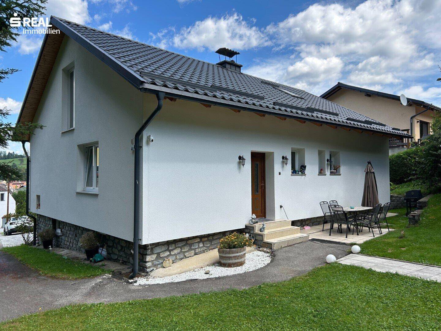 Seitenansicht des Hauses mit kleiner Terrasse, Gartenmöbeln und gepflegtem Rasenbereich.