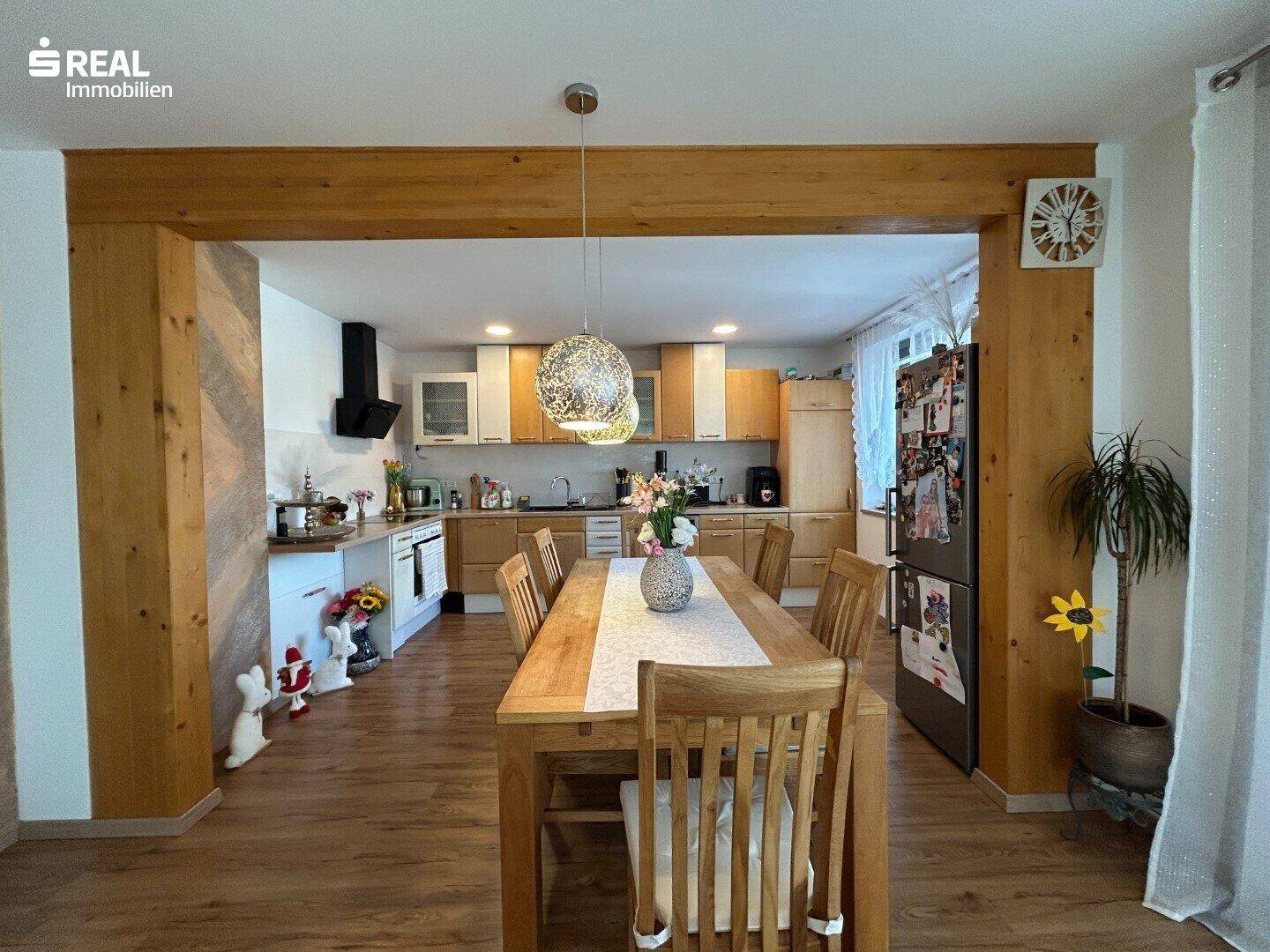 Offene Küche mit Essbereich, Holztisch und modernen Einbauschränken unter sichtbaren Holzbalken.