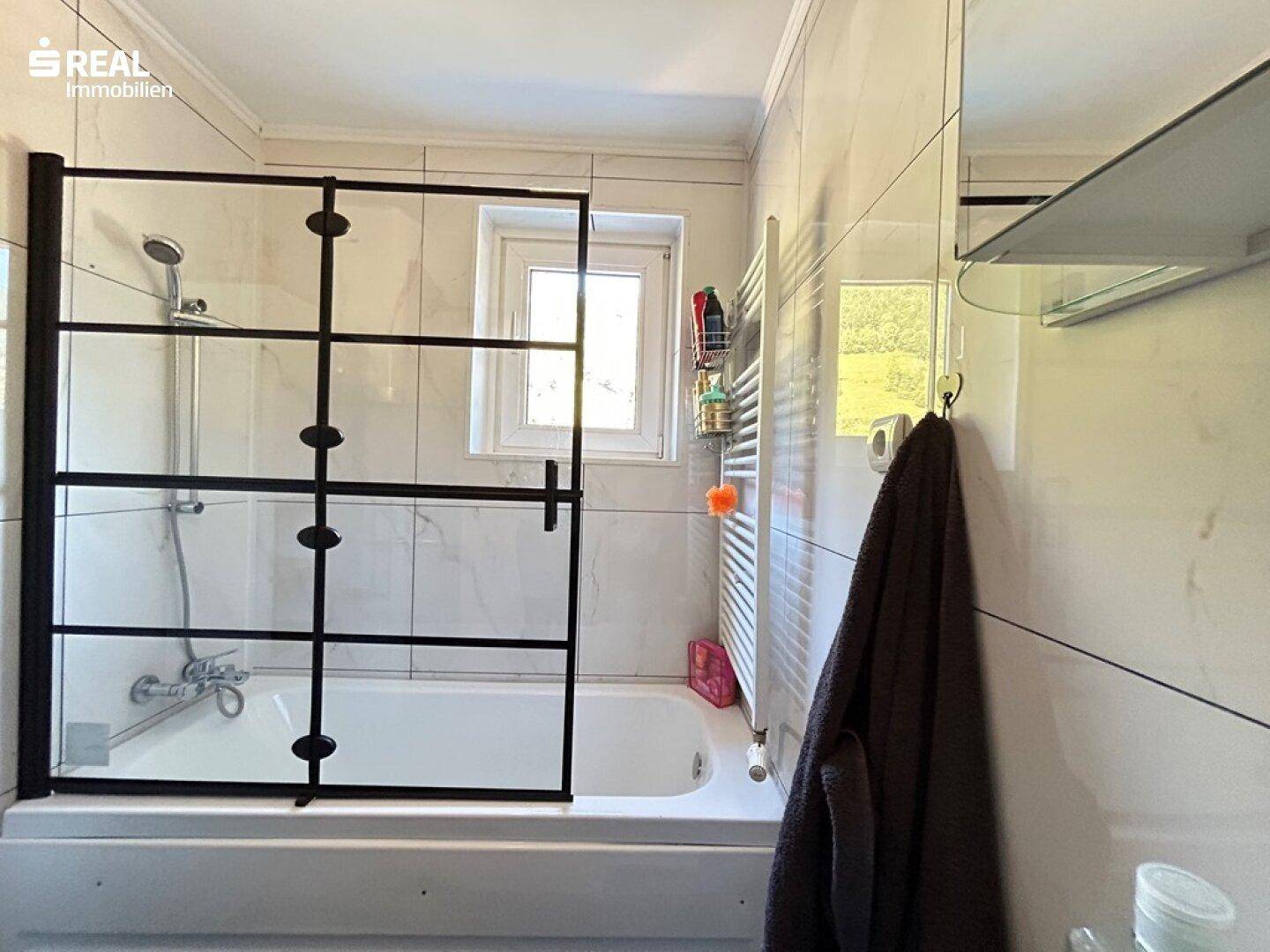 Helles Badezimmer mit Badewanne, Duschabtrennung und beheiztem Handtuchhalter am Fenster.