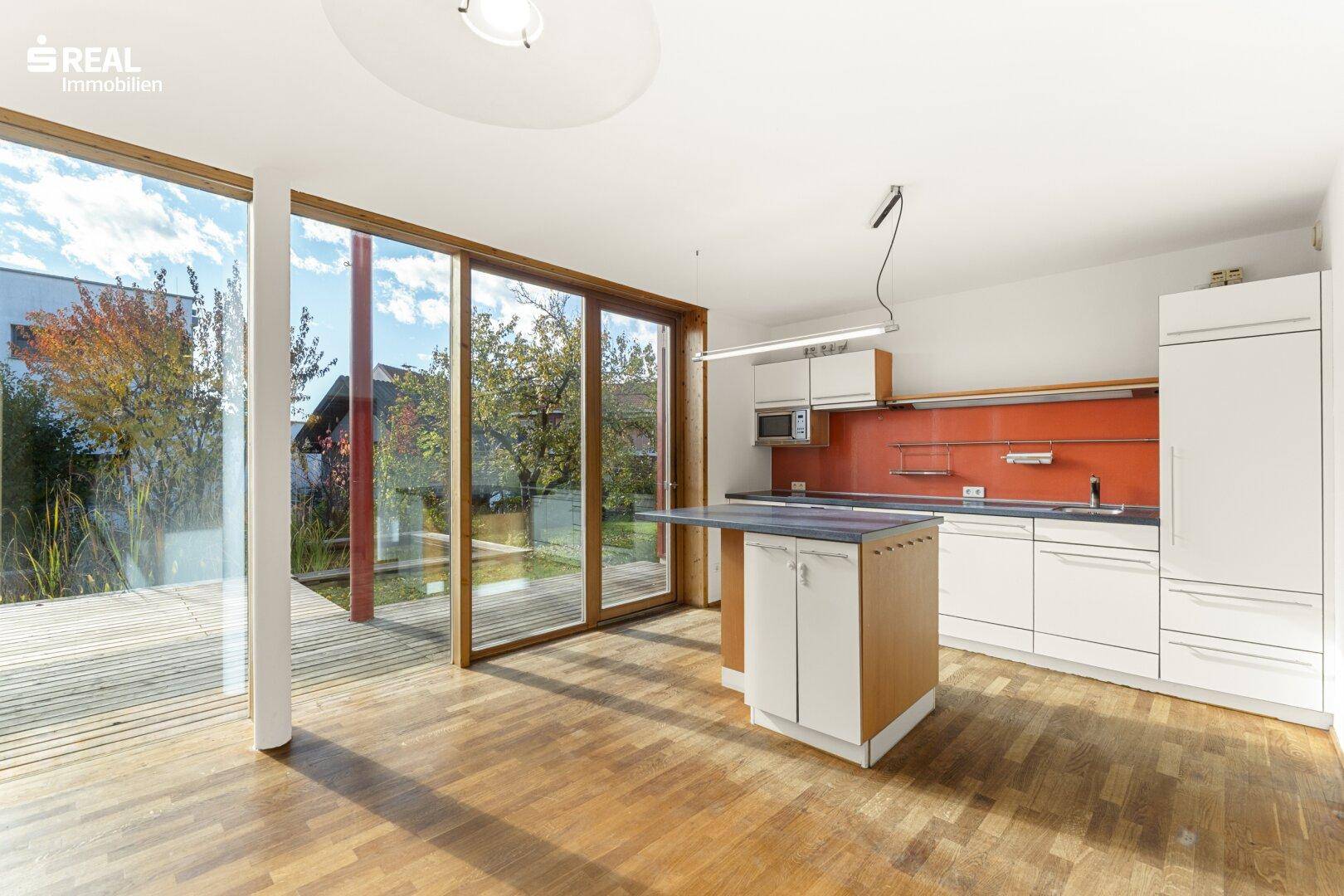 Offene Küche mit weißen Schränken, roter Rückwand und Zugang zur Holzterrasse.