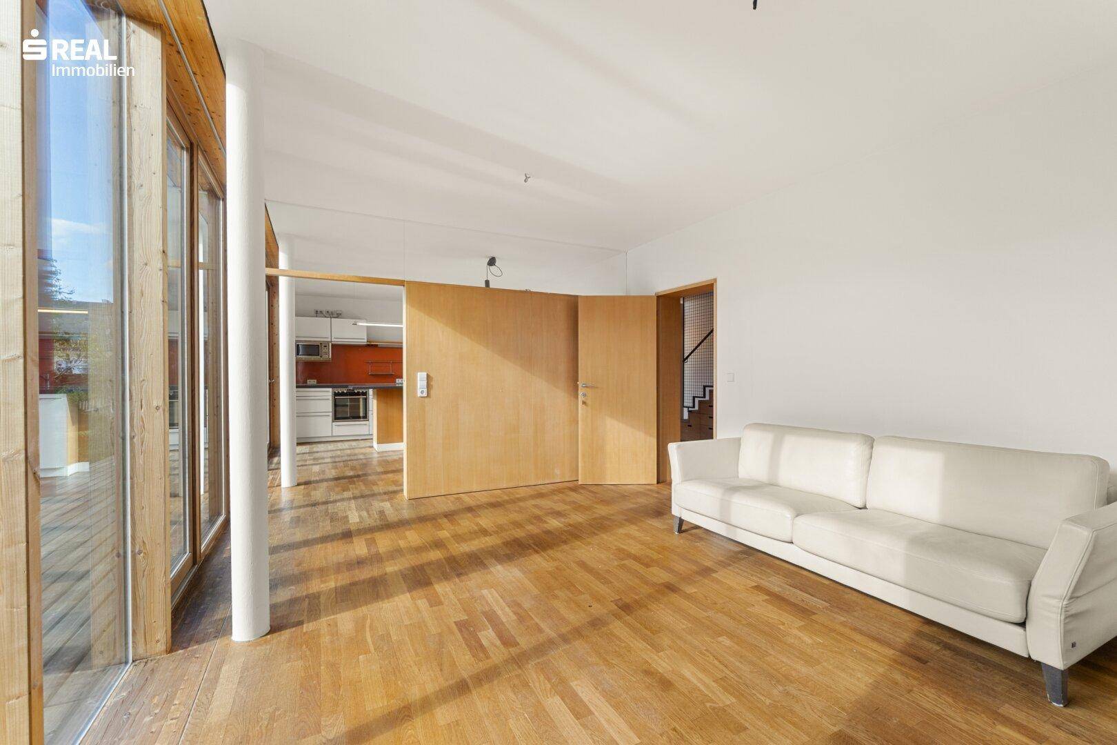 Helles, leeres Wohnzimmer mit Holzboden und großen Fensterfronten, die viel Tageslicht hereinlassen.