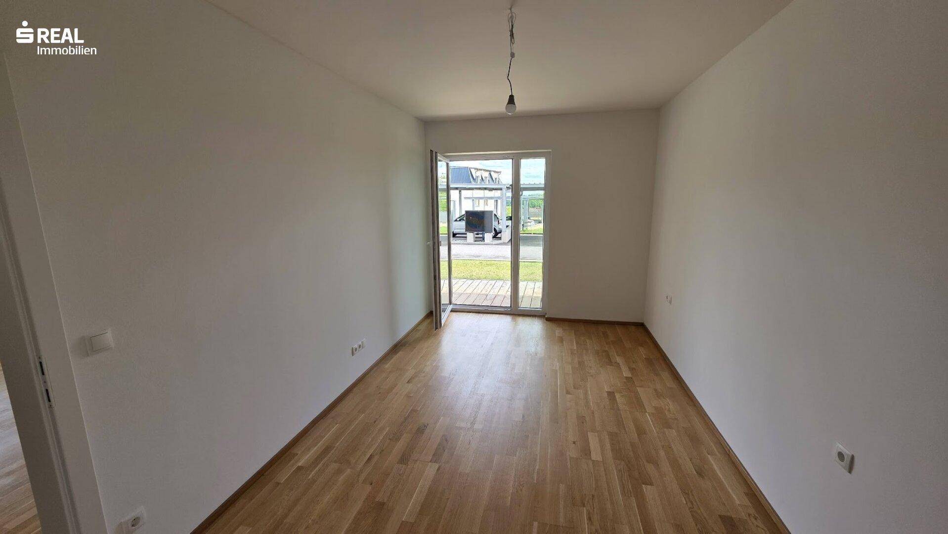 Leerer Raum mit Holzboden und großer Glastür, die viel Tageslicht hereinlässt.