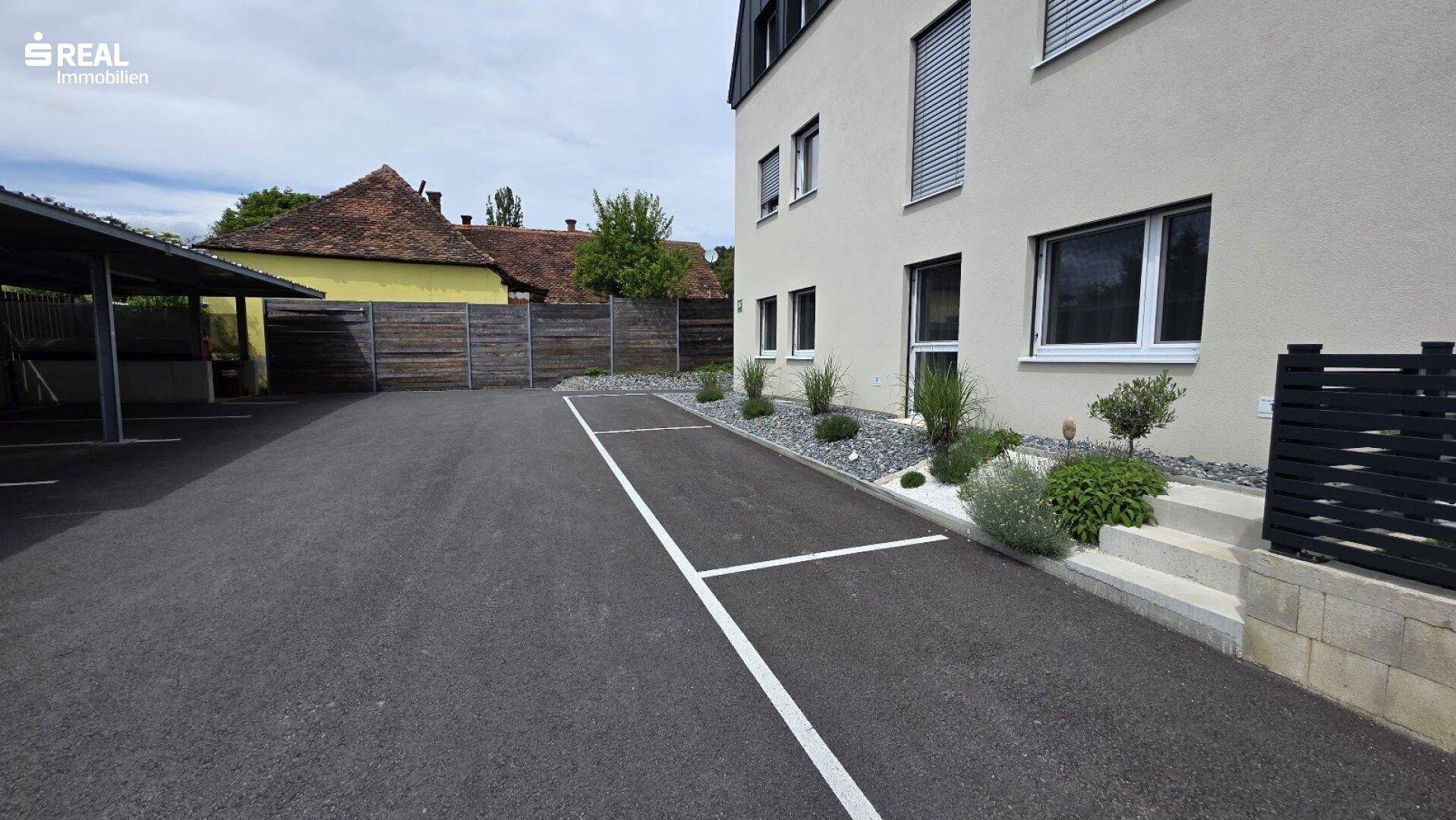 Außenbereich mit asphaltiertem Parkplatz, Carport und kleiner Bepflanzung am Gebäude.