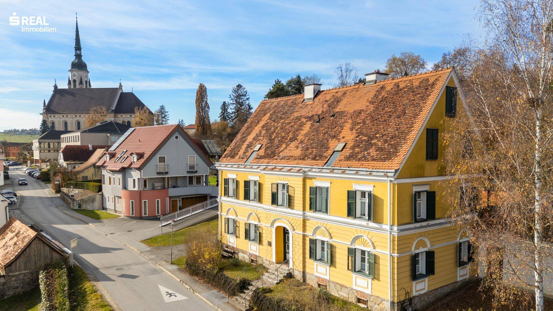 Historisches gelbes Gebäude mit grünen Fensterläden und Blick auf die nahegelegene Kirche.