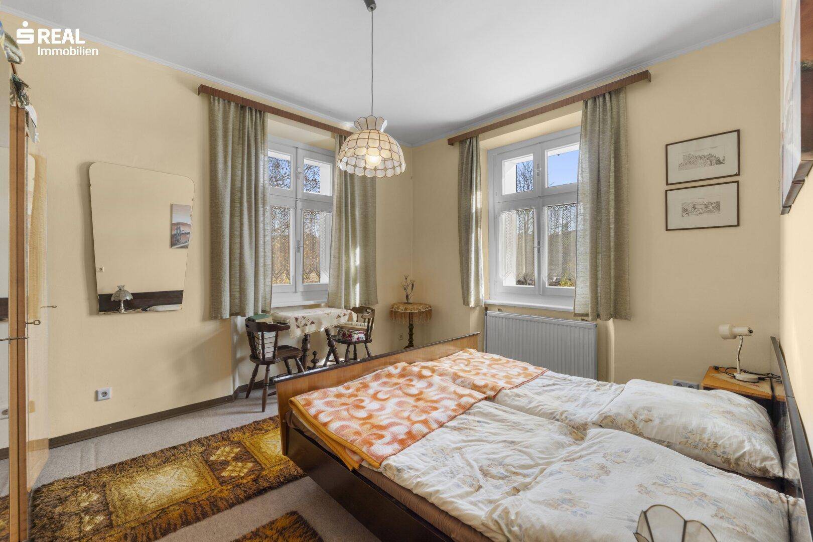Helles Schlafzimmer mit Doppelbett, einfacher Einrichtung und zwei großen Fenstern.
