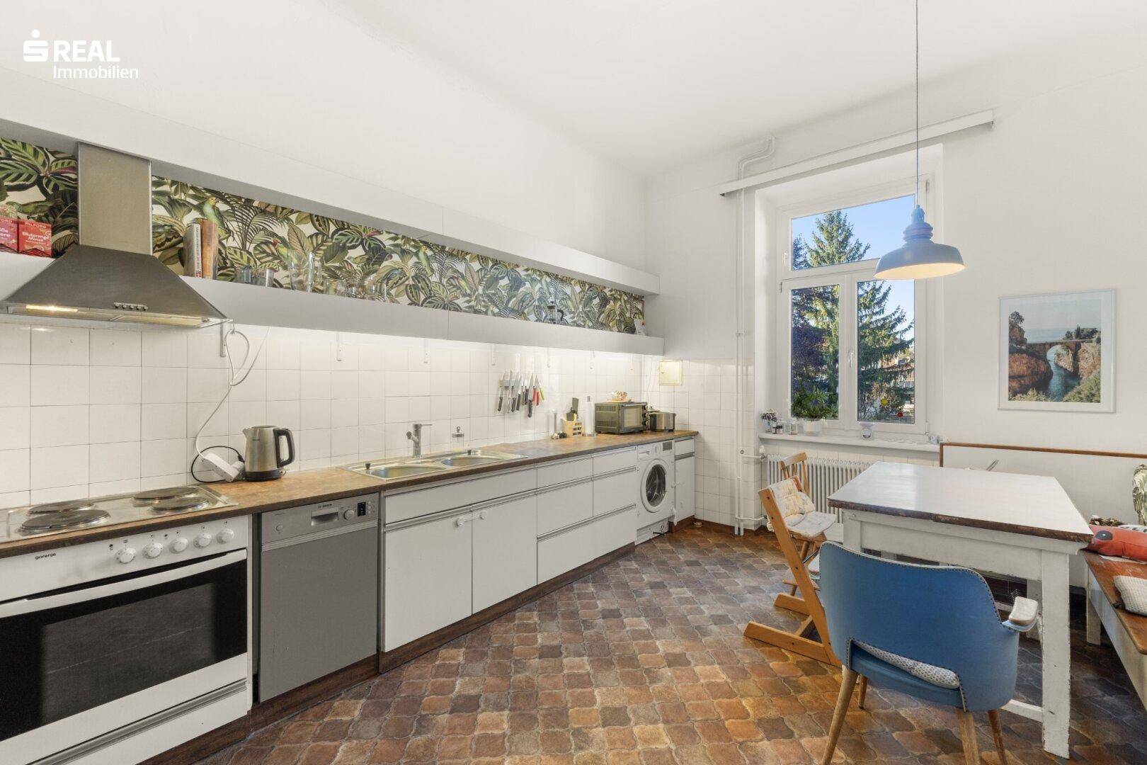 Funktionale Küche mit weißen Schränken, Essplatz und Fenster mit Blick ins Freie.