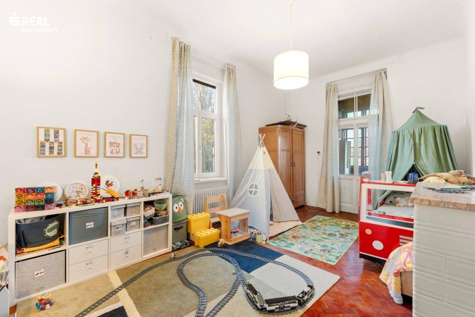 Farbenfrohes Kinderzimmer mit Spielteppich, viel Stauraum und hohen Fenstern.