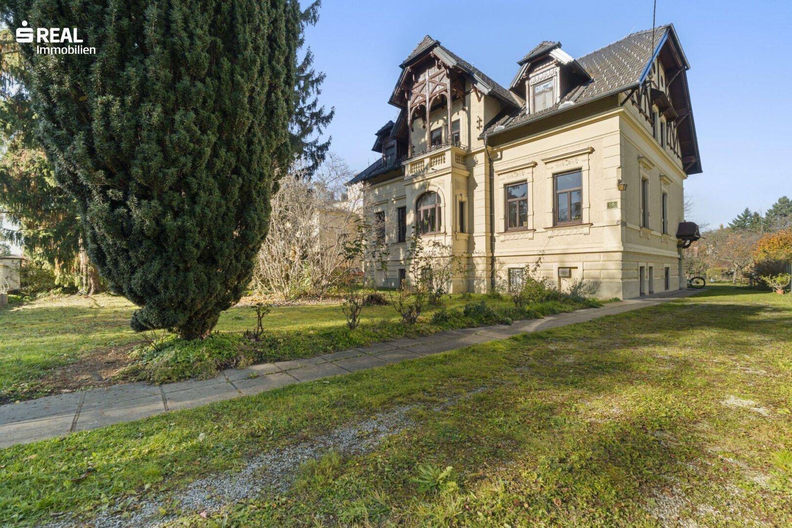 Elegante Villa mit gepflegtem Vorgarten und altem Baumbestand unter blauem Himmel.