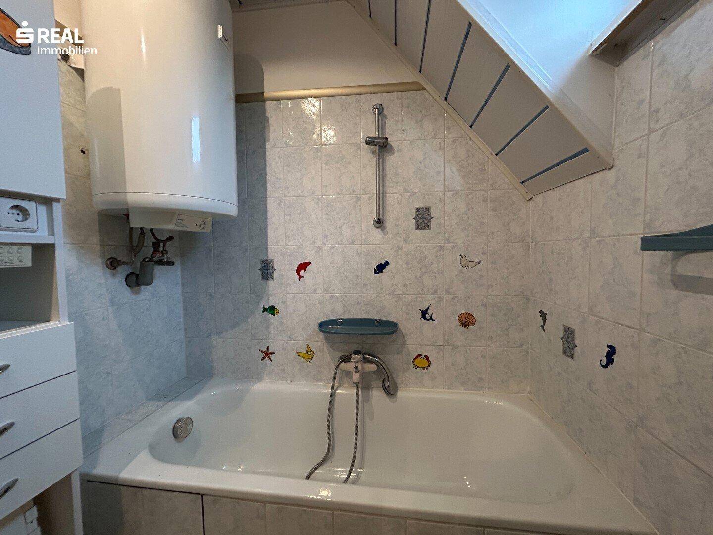 Badezimmer mit Badewanne, Duschvorrichtung und Wandfliesen mit maritimen Motiven.