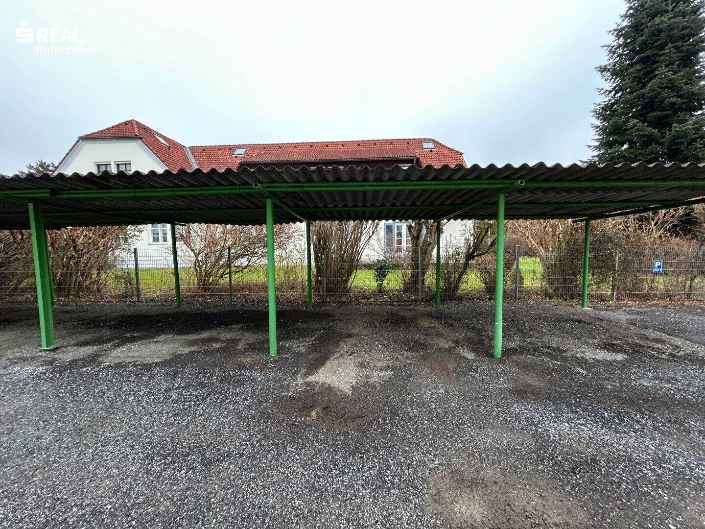 Überdachter Parkplatz oder Carport mit grün gestrichenen Metallstützen und Kiesboden.