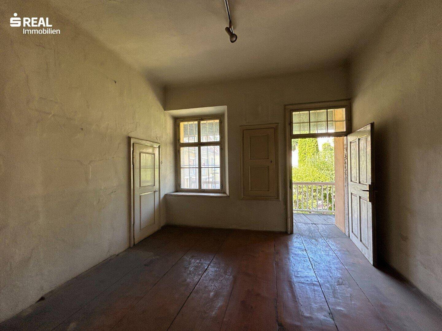 Raum mit Holzdielenboden, zwei Fenstern und Zugang zu einem Balkon, der eine Renovierung erfordert.