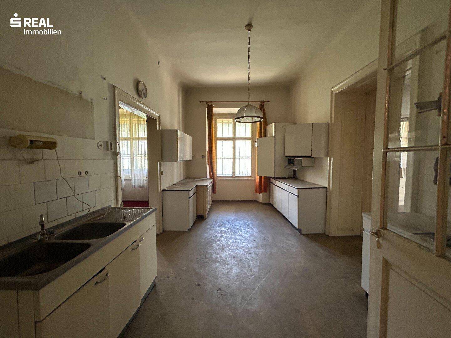 Alte Küche mit einfachen Schränken, Doppelwaschbecken und einem Fenster, das renovierungsbedürftig ist.