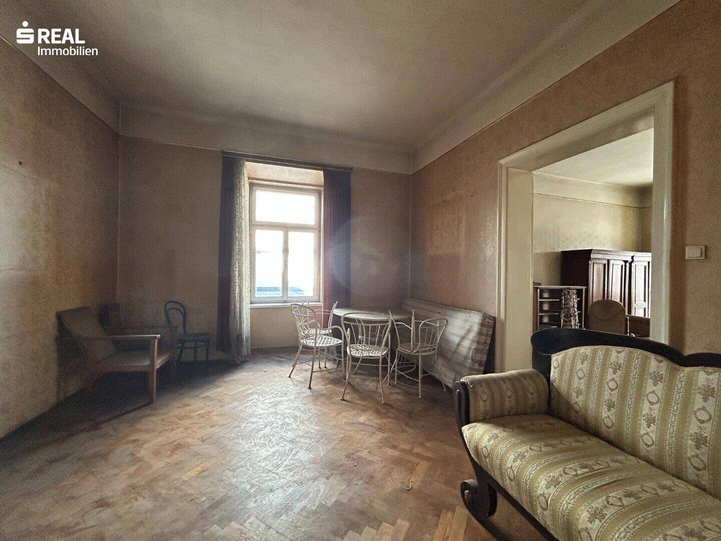 Wohnzimmer mit Fischgrätparkett, einem Sofa und einem kleinen Tisch am Fenster, das renovierungsbedürftig ist.