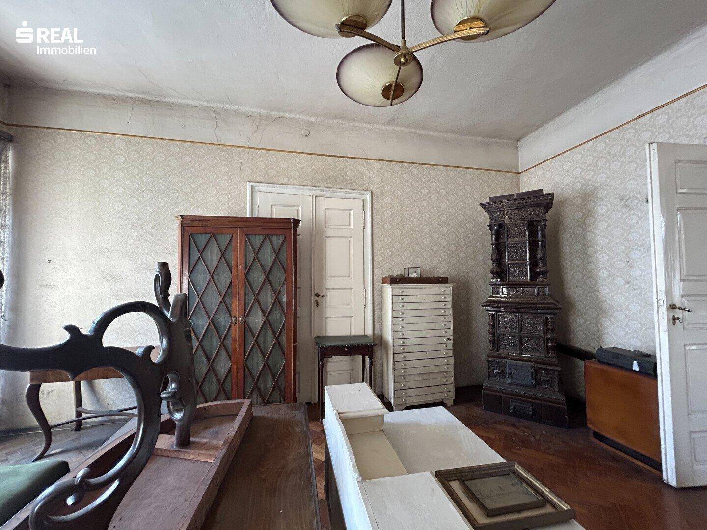 Geräumiges Zimmer mit antiken Möbeln, gemusterter Tapete und einem traditionellen Kachelofen.