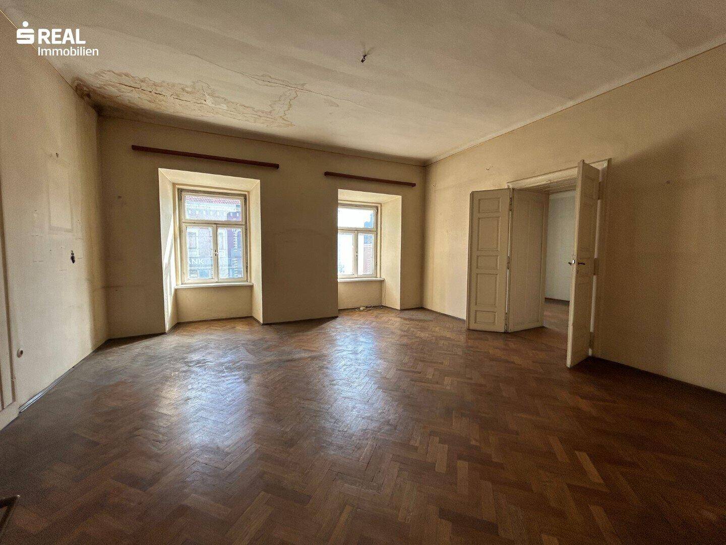Heller Raum mit Fischgrätparkett und zwei Fenstern, die viel Tageslicht hereinlassen, renovierungsbedürftig.