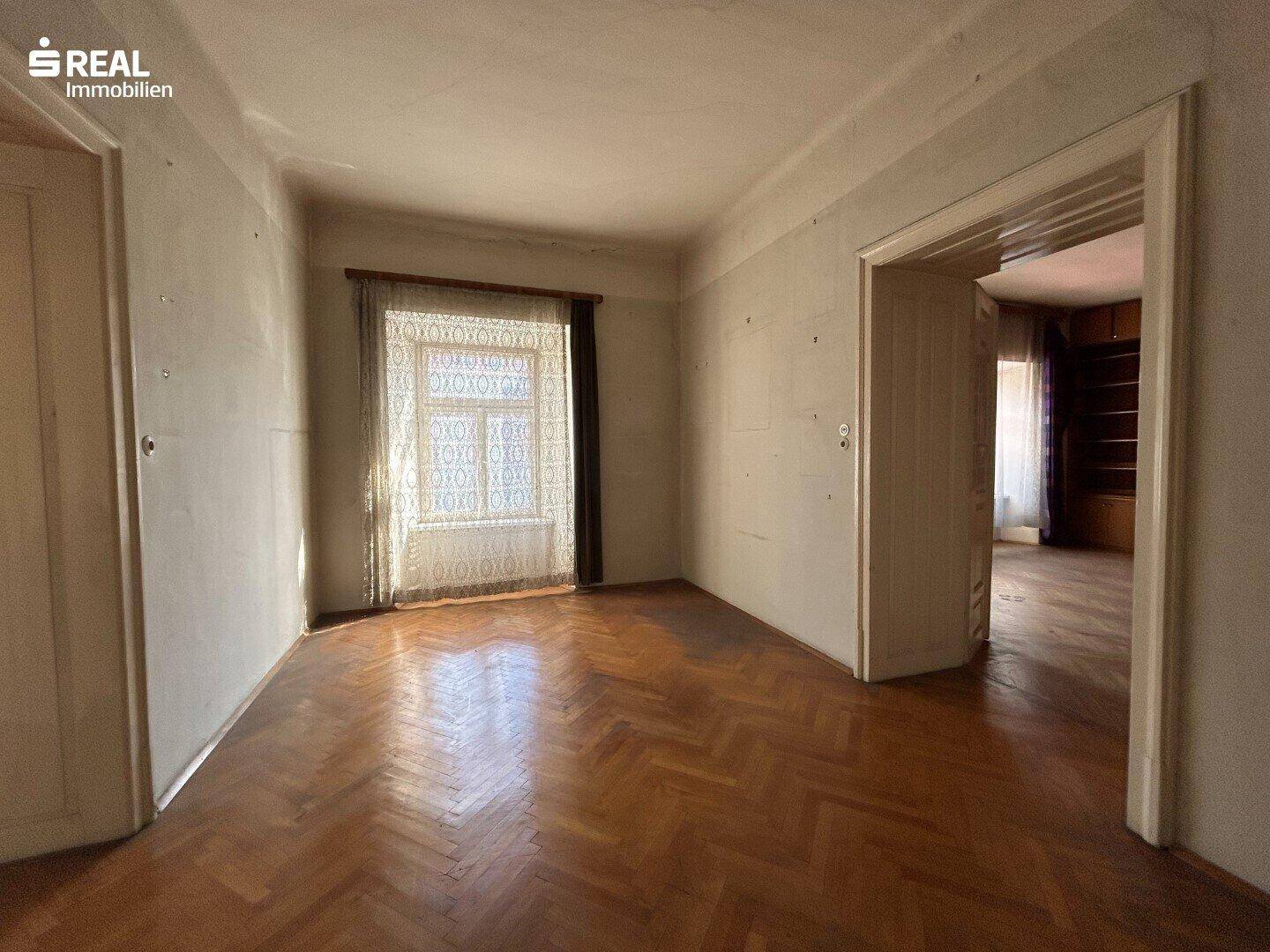 Leerer Raum mit Fischgrätparkett und einem Fenster, das Tageslicht hereinlässt, ideal für eine Renovierung.