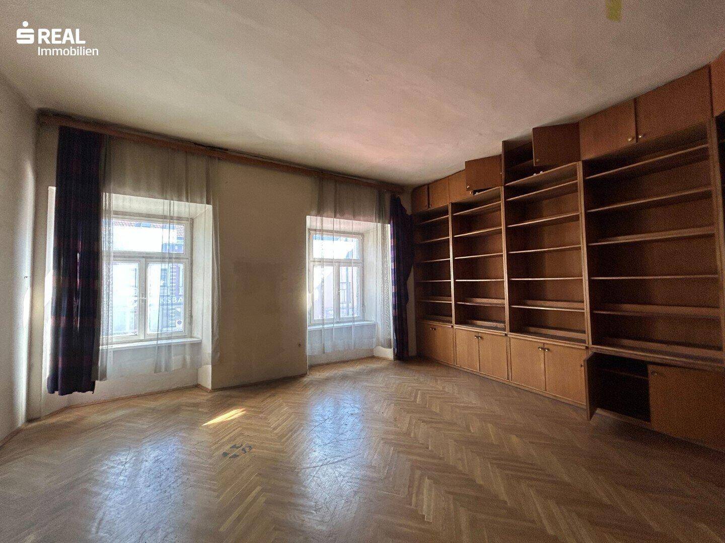 Geräumiger Raum mit Fischgrätparkett, eingebauten Holzregalen und zwei Fenstern, die für Helligkeit sorgen.