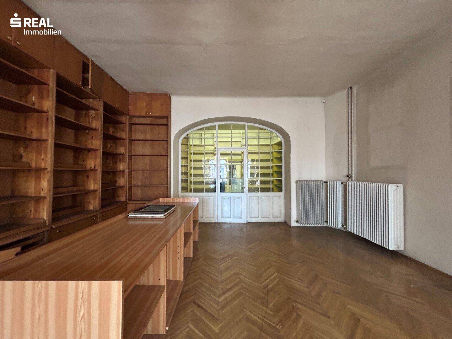 Ehemaliger Ladenraum mit Holzregalen und einem großen Bogenfenster zur Straße, renovierungsbedürftig.