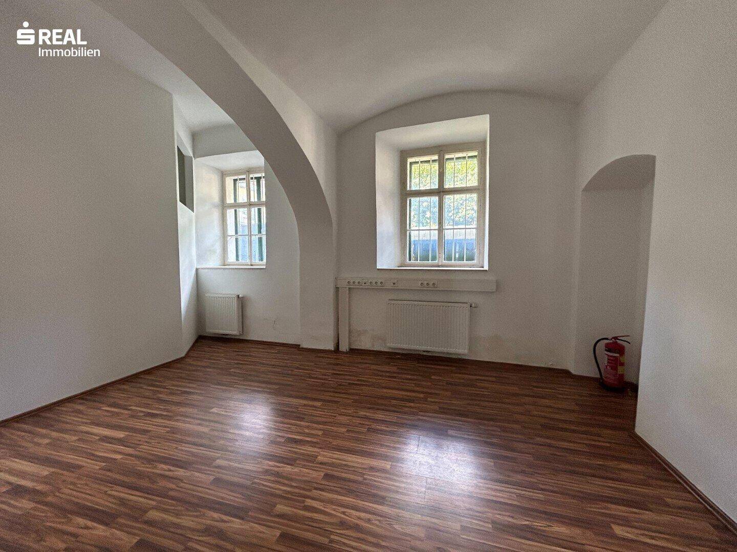 Geräumiger Ladenraum mit Laminatboden, gewölbter Decke und zwei Fenstern, die viel Licht hereinlassen.