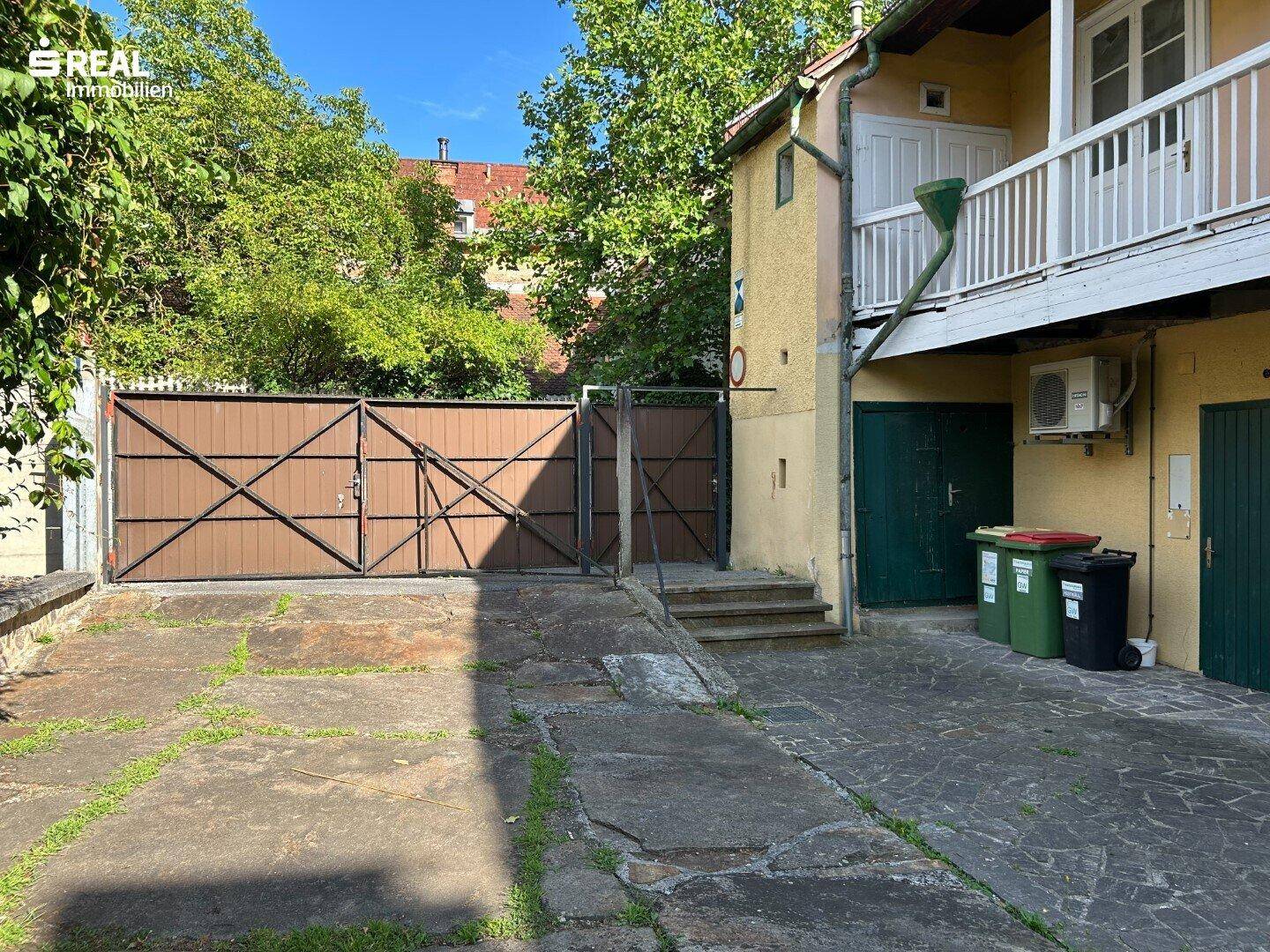 Gepflasterter Hofbereich mit großen Holztoren und Mülltonnen, der als Zufahrt dient.