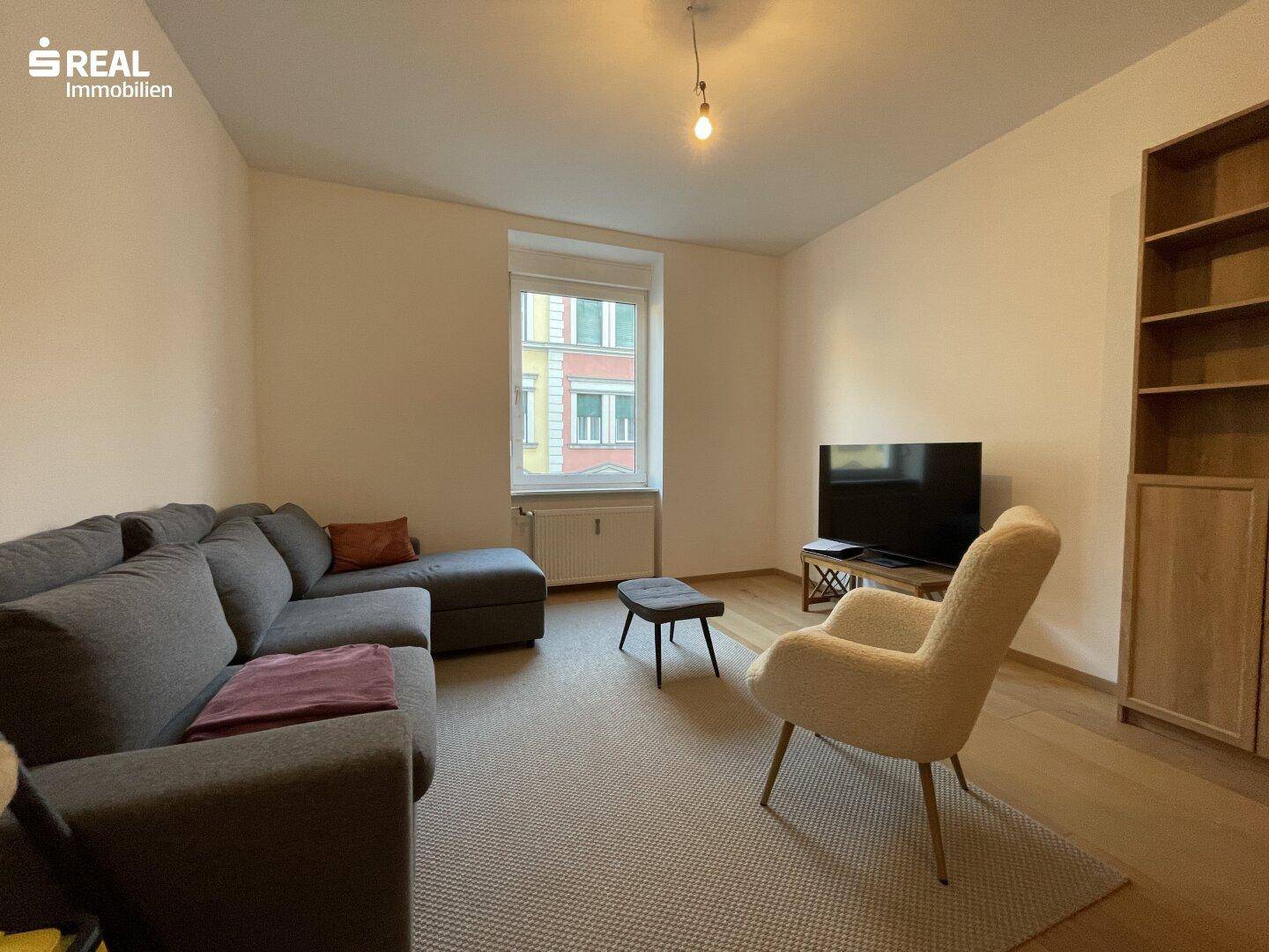 Gemütliches Wohnzimmer mit grauem Ecksofa, Sessel und Blick auf die Straße.