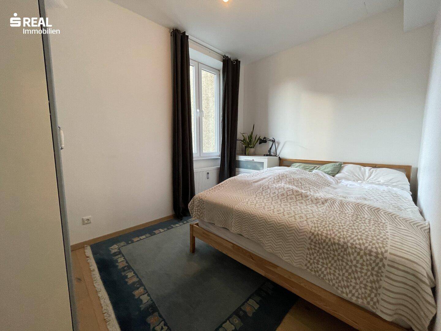 Schlafzimmer mit Holzbett, gemusterter Bettwäsche und Fenster mit dunklen Vorhängen.