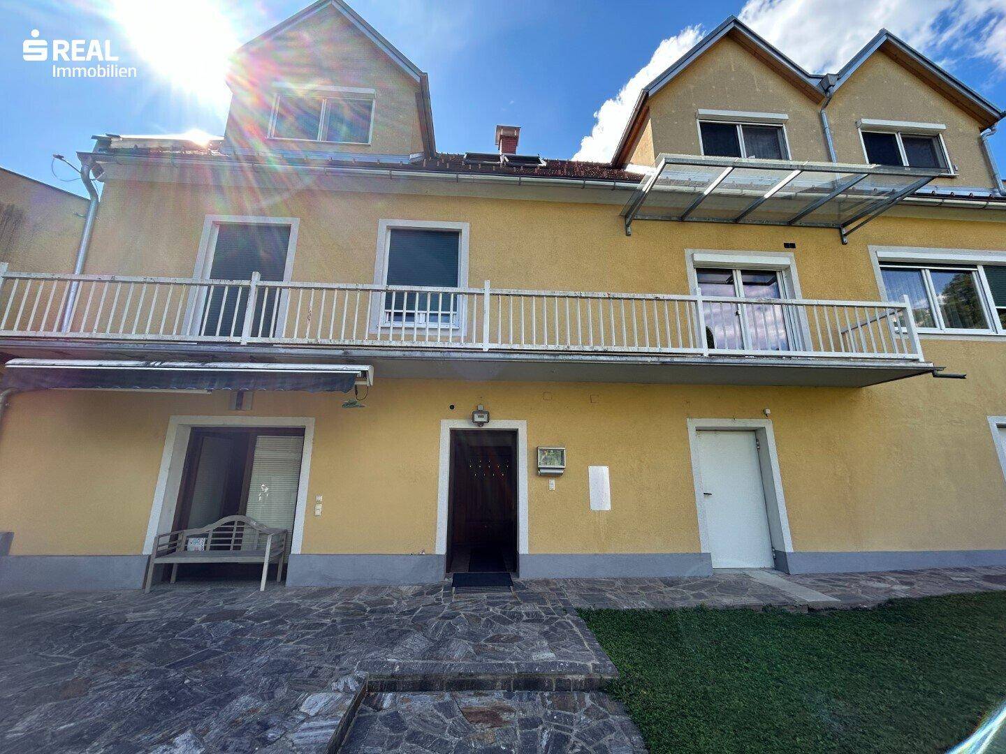 Gelbe Fassade eines Hauses mit Balkon und Eingangsbereich unter strahlendem Himmel.