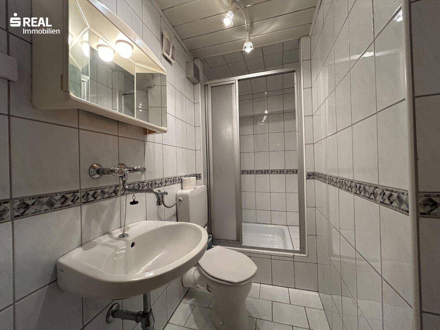Kleines Badezimmer mit weißen Fliesen, Dusche und Waschbecken, funktional eingerichtet.