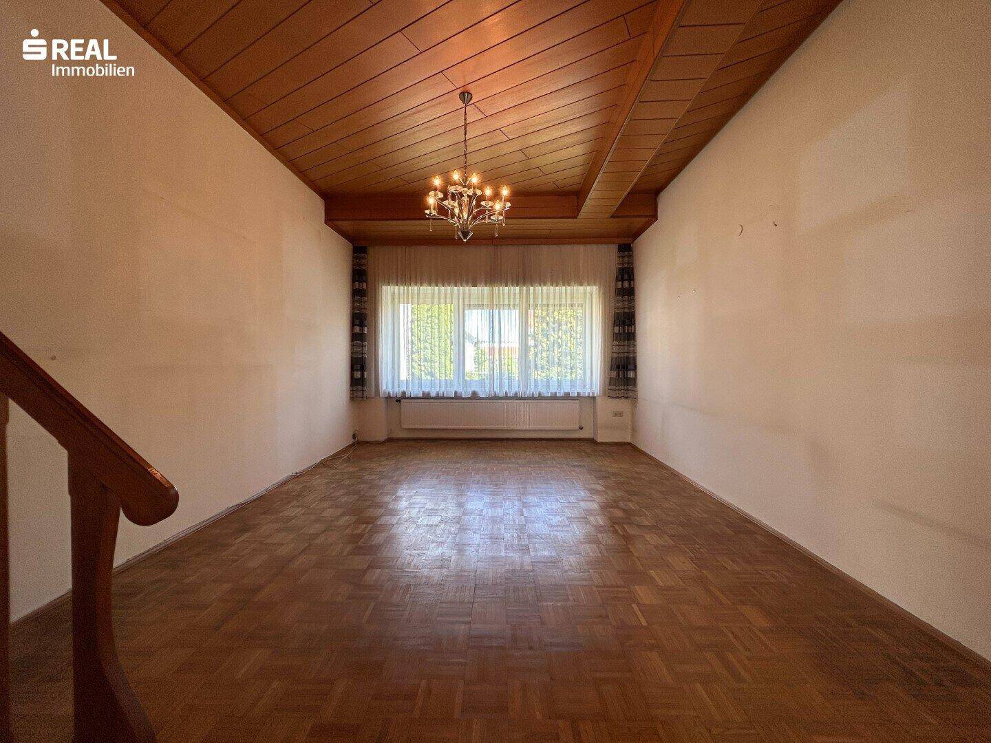 Geräumiges Wohnzimmer mit Parkettboden, Holzdecke und großem Fenster, klassisch eingerichtet.