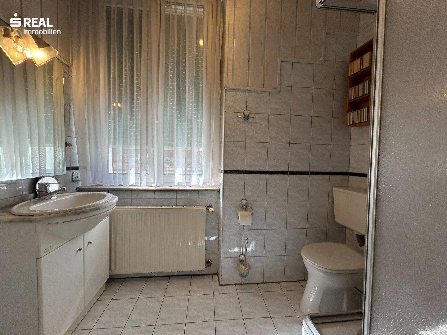 Badezimmer mit Dusche, Waschbecken und Fenster, hell gefliest und funktional ausgestattet.